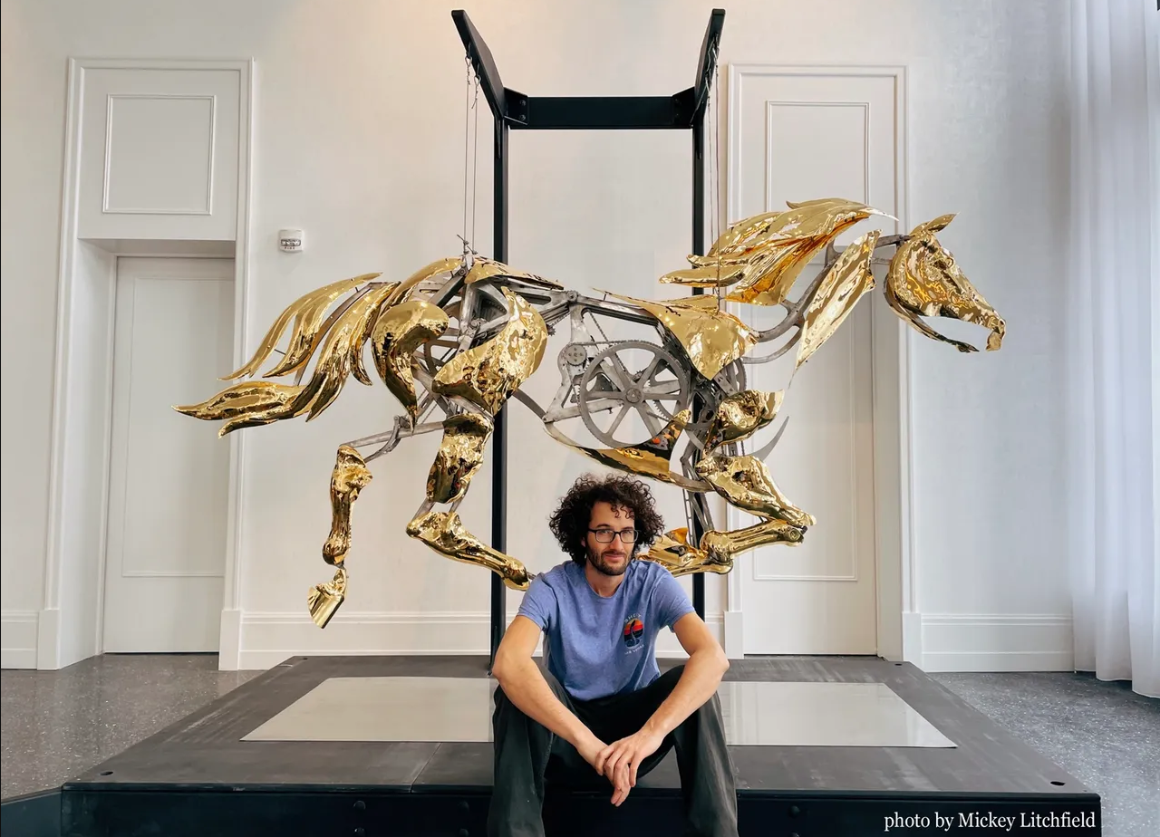 Artist sits on ground in front of large metal, mechanical horse statue with gears exposed.