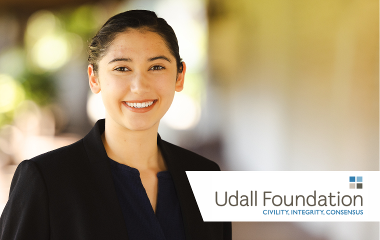 Young woman with black hair and black button up dress shirt stands on tile walkway with adobe brick wall on Santa Clara's campus. A graphic element reads "Udall Foundation - Civility, Integrity, Consensus"
