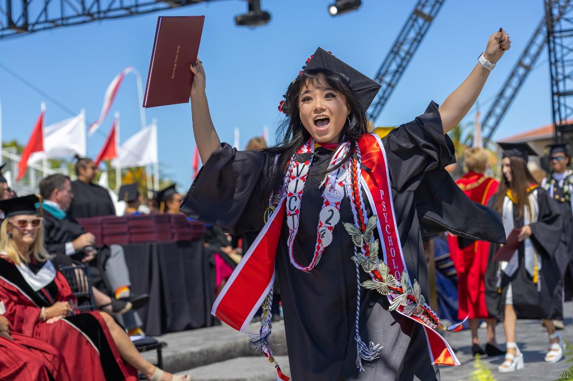 Woman student in a graduation gown and mortar board celebrates with her hands over her head, holding diploma on stage.