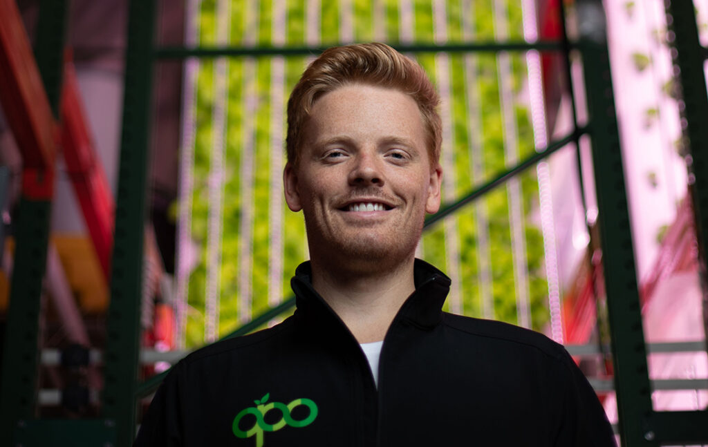 A young man with stylized red hair, wearing a black zip up jacket stands in front of a vertical wall of plants.