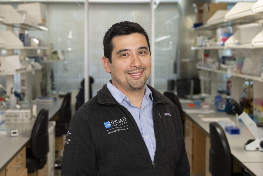A man with dark hair and a black zip up jacket over a blue collared shirt stands between two rows of tables in a lab.