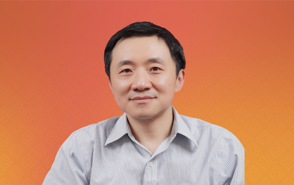 A man with dark hair and a light colored button up shirt sits in front of an orange background.