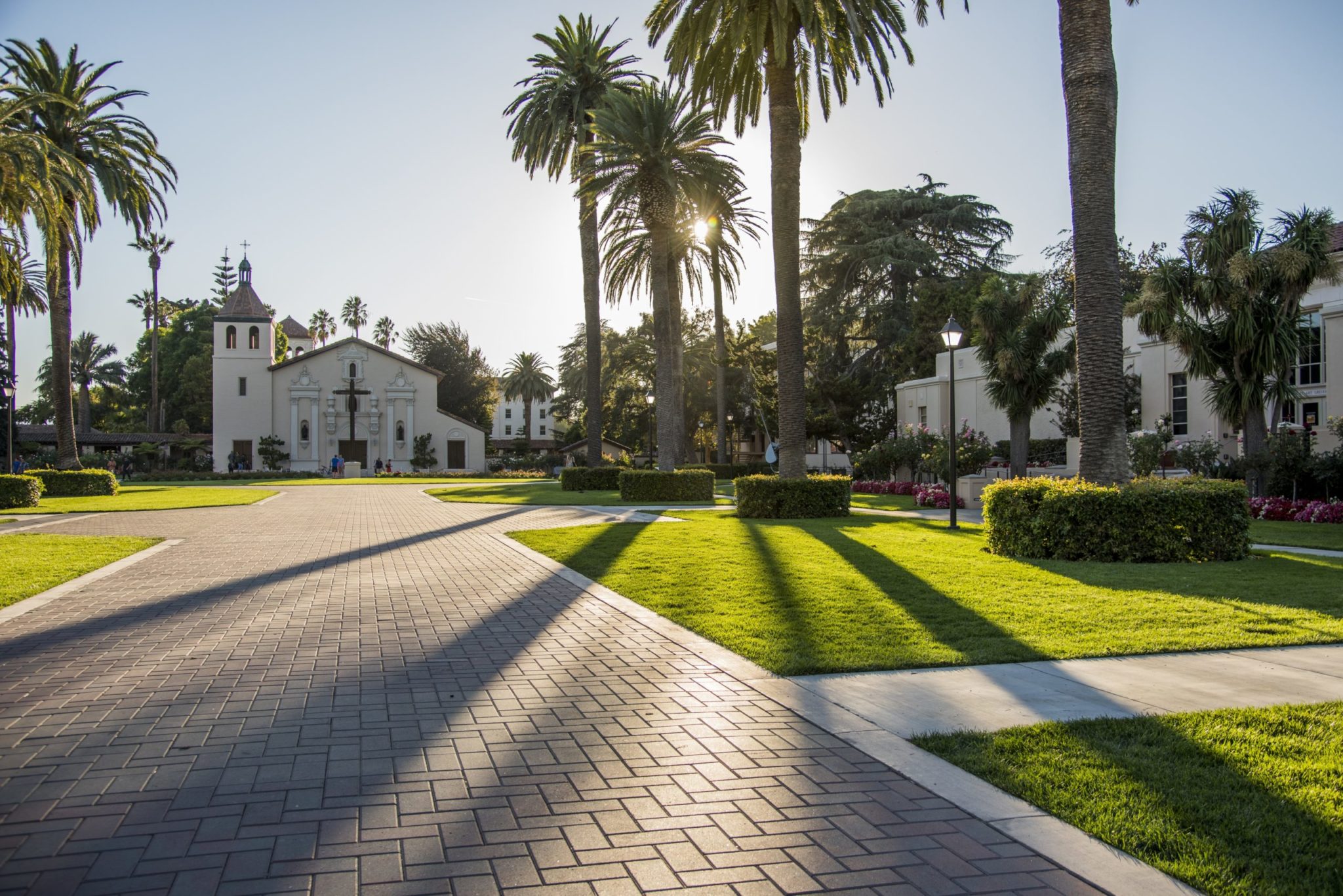 A photo of the sidewalk leading up to the Mission Church with sun peeking over the trees and casting shadows onto the walkway.