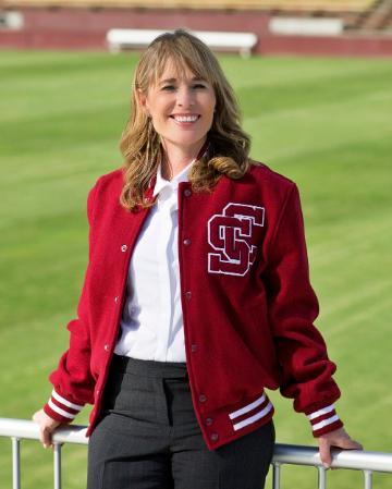 Woman in Santa Clara letteman jacket, leaning against a steel rail with a green grass field in the background.