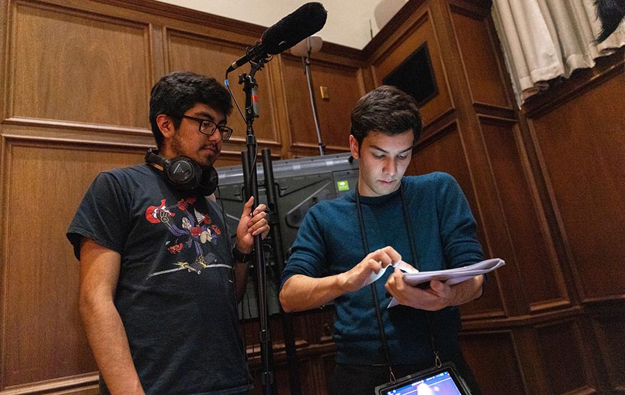 Two college aged students huddle in the corner of a wood paneled room. They are holding filming equipment including a boom mike and notepad.