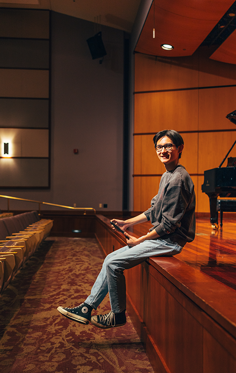 A young man sits on the edge of an empty recital halls stage looking to the camera, holding an iPad in his hand, with a piano in the background.