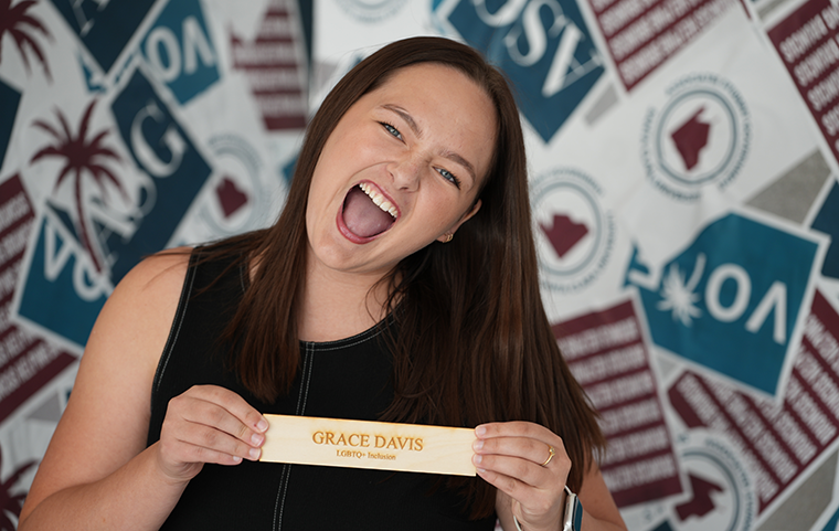 A young woman with a wide cheering smile holds her senator placard which says Grace Davis and LGBTQ+ Inclusion.