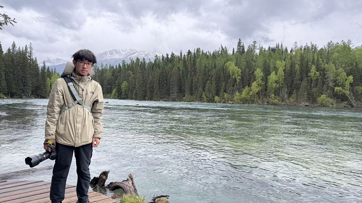 A young man holding a telephoto camera stands next to a crystal clear river, and a dense forest and snow-capped mountains in the background.