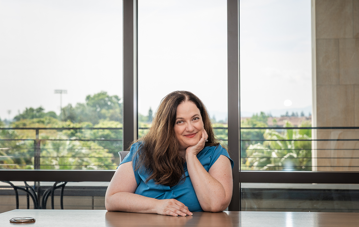 A woman in a blue blouse sits at a conference room table, one arm resting casually on the tabletop while she leans and rests her head on the palm of her propped arm. A view of the sky is visible through the rooms dramatic, large windows.