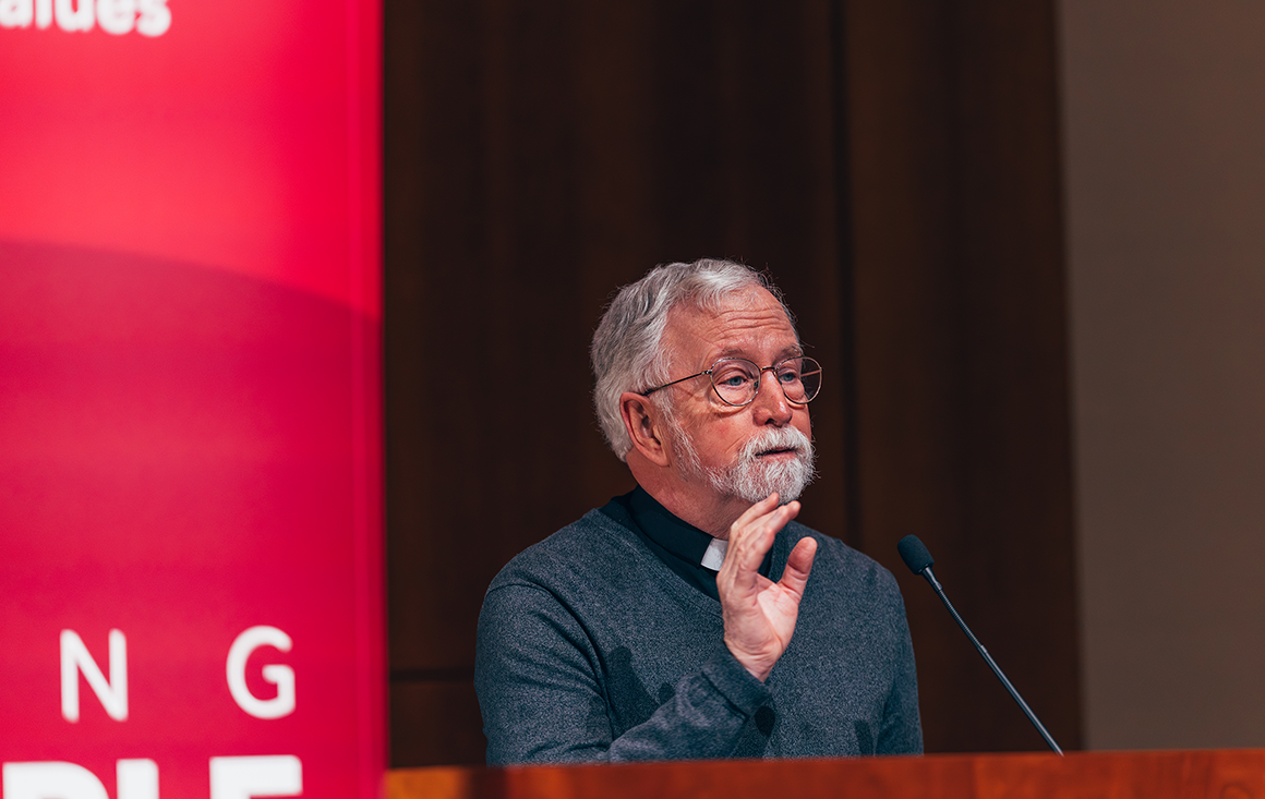 An older white Jesuit priest orates at a podium.