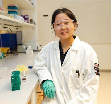 A female student wearing a lab coat and goggles at a counter in a chemistry lab.