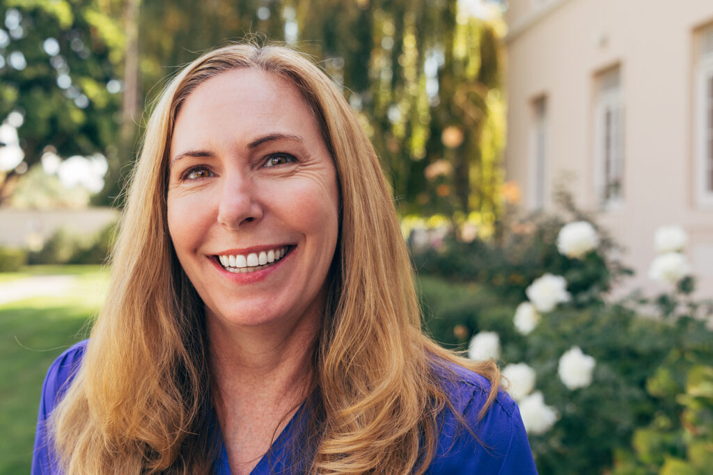 A blonde woman in a blue blouse smiles warmly, standing among white roses and a willow tree.