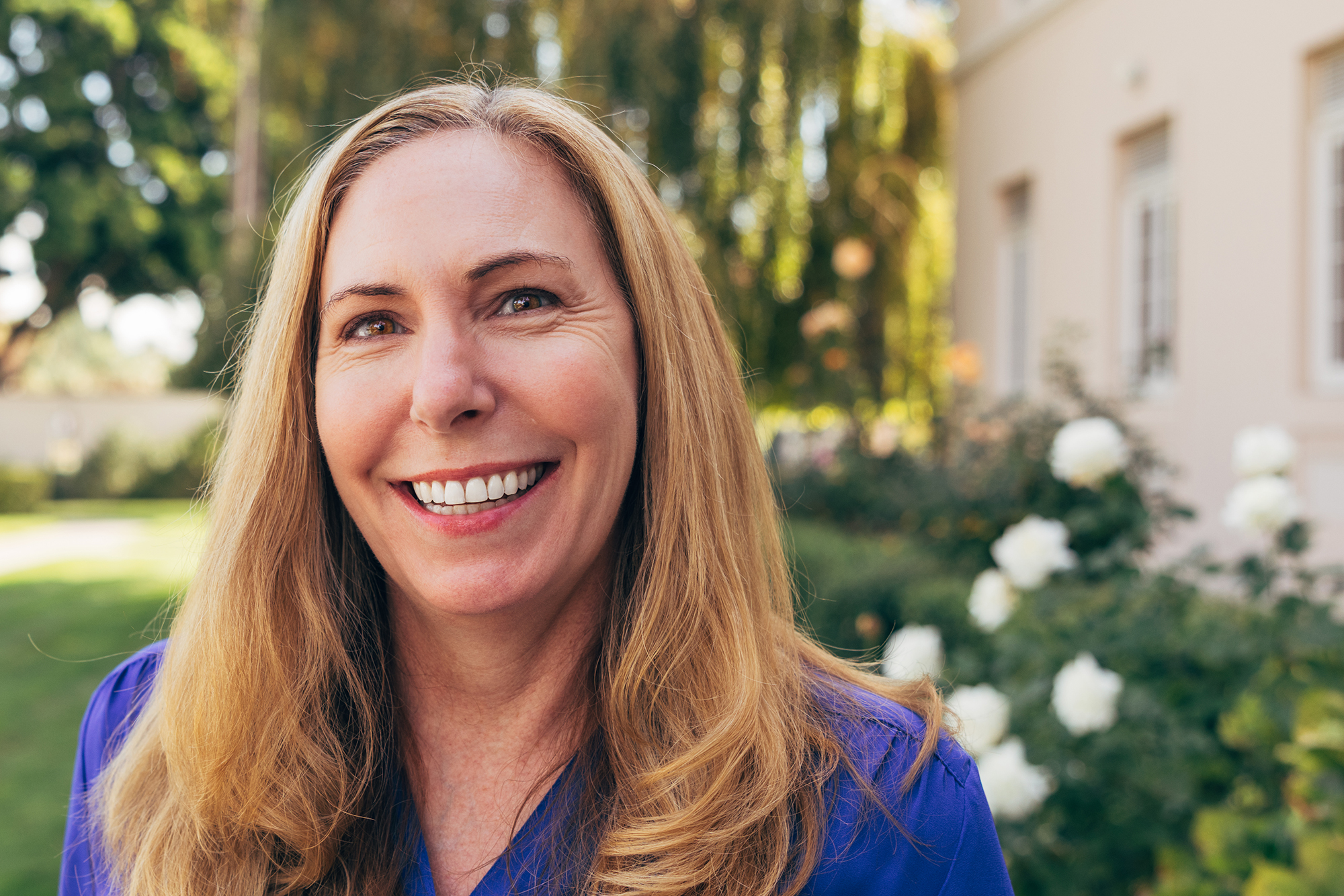 A blonde woman in a blue blouse smiles warmly, standing among white roses and a willow tree.