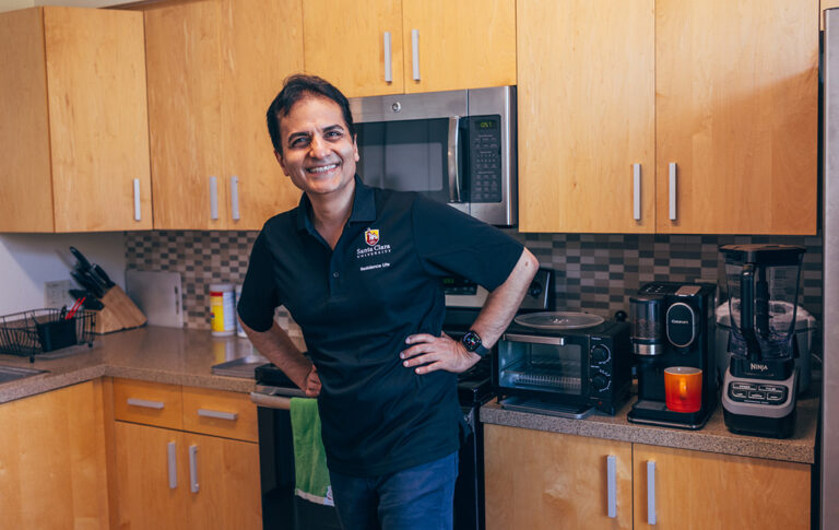 A older Persian man stands with his hands playfully on his hips and a beaming smile in a kitchen, while brewing coffee.