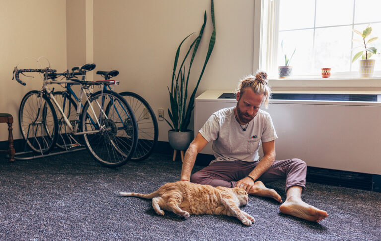 A blonde, white man pets his cat in a carpeted dorm room, surrounded by bikes and plants.
