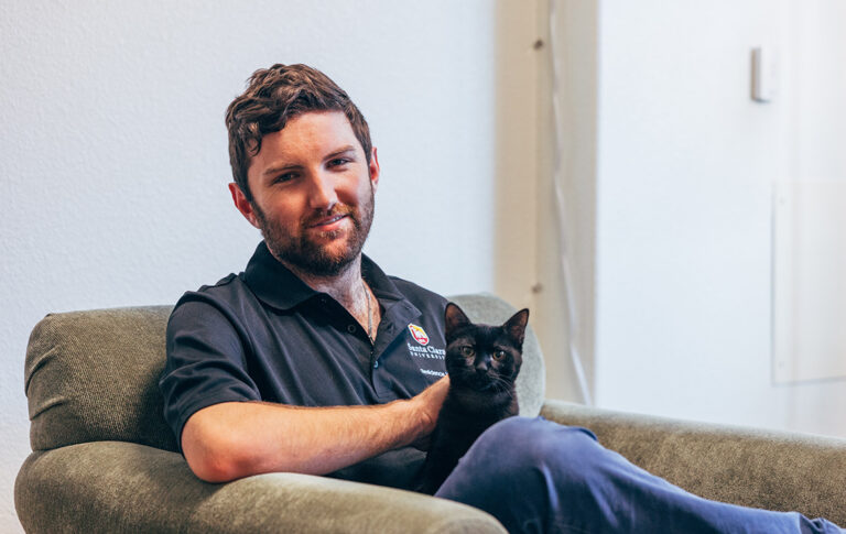 A young white man sits on a couch, petting a black cat.
