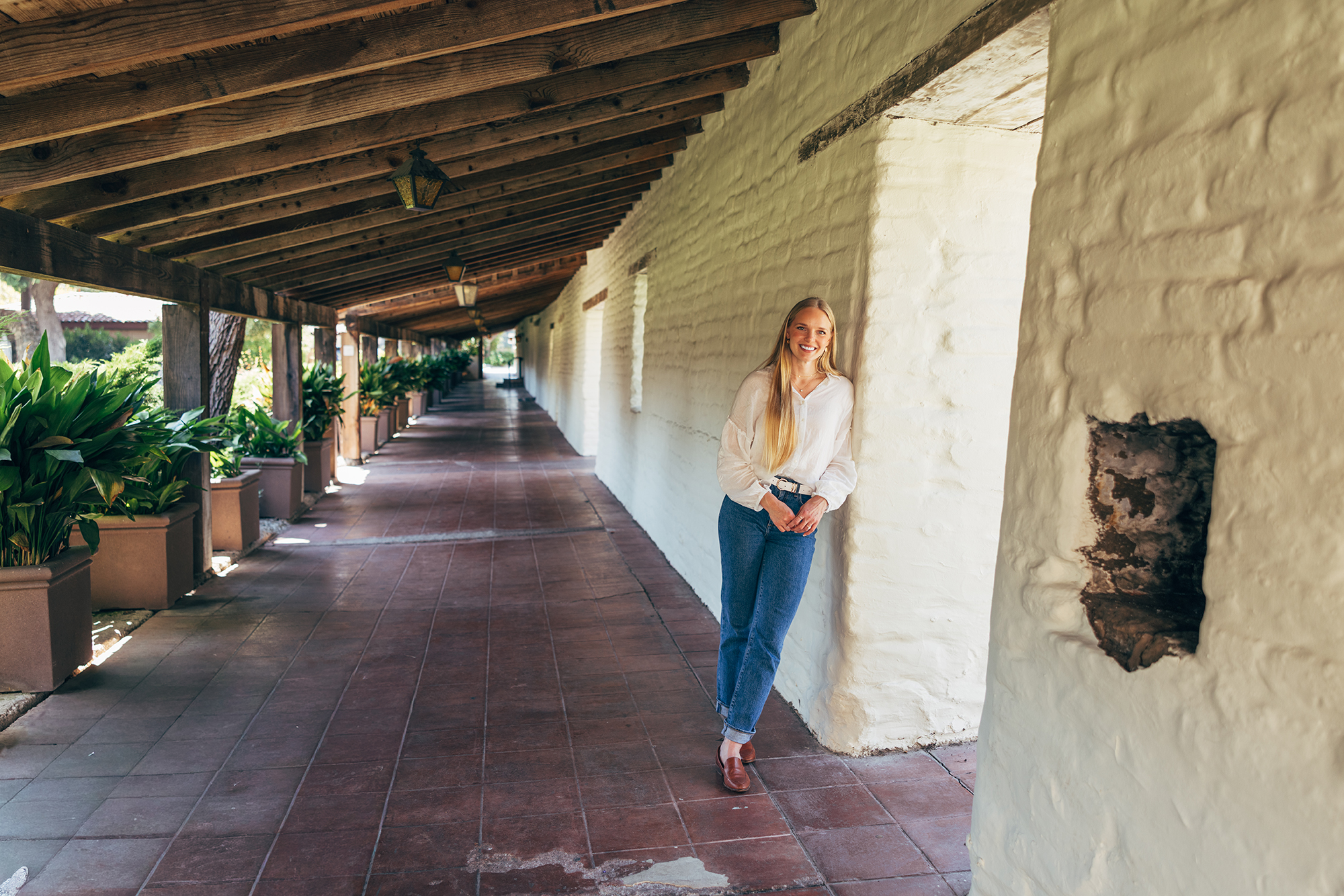 Woman in white long sleeved shirt and jeans leans against Adobe brick on walkway next to Adobe Lodge on Santa Clara University campus.