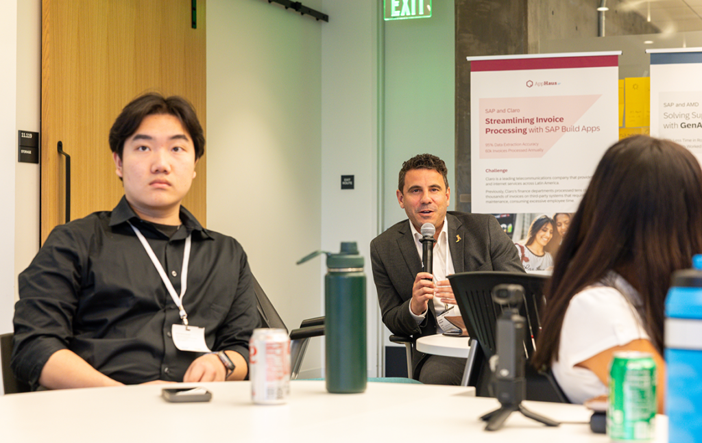 Man wearing suit speaks into microphone while sitting at a table. Two students are sitting at a different table in the foreground.