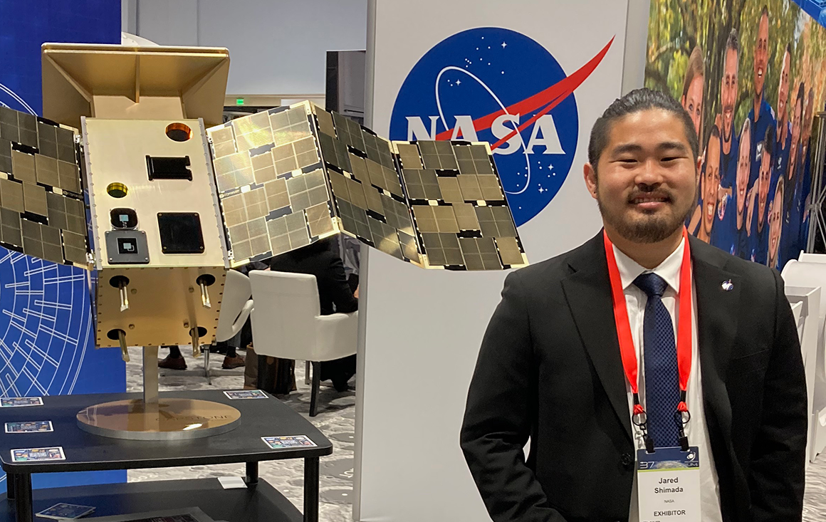 Man in suit stands in front of a wall with a NASA logo and a table with a gold plated piece of machinery that resembles a small satellite.