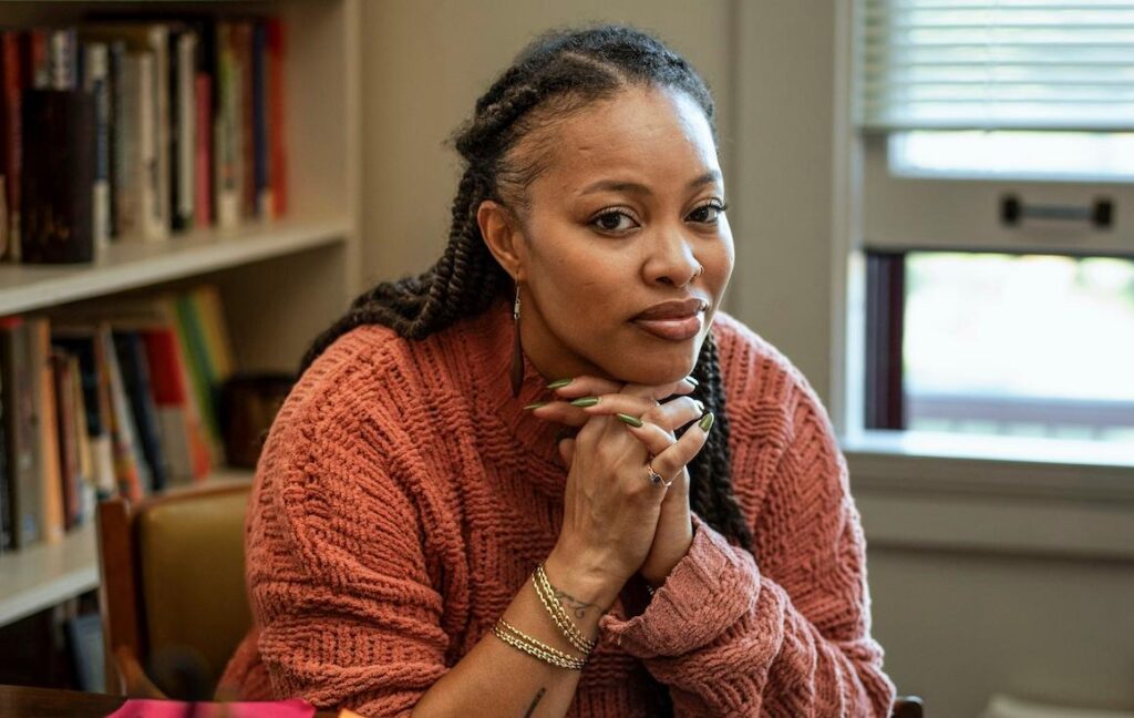 A woman with braids sits in an academic office with her hands clasped just under her chin.