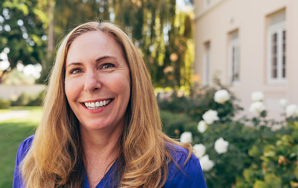 A blonde woman in a blue blouse smiles warmly, standing among white roses and a willow tree.
