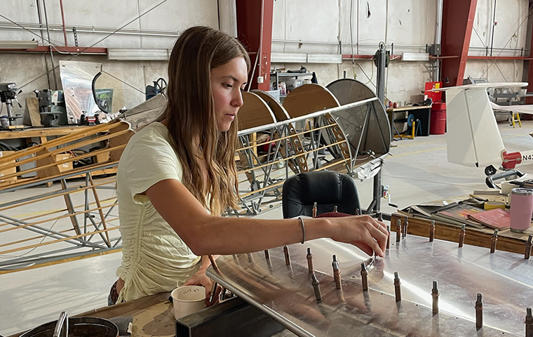 Woman in white shirt sits at a table working on a piece of airplane equipment with a deconstructed airplane in the background.