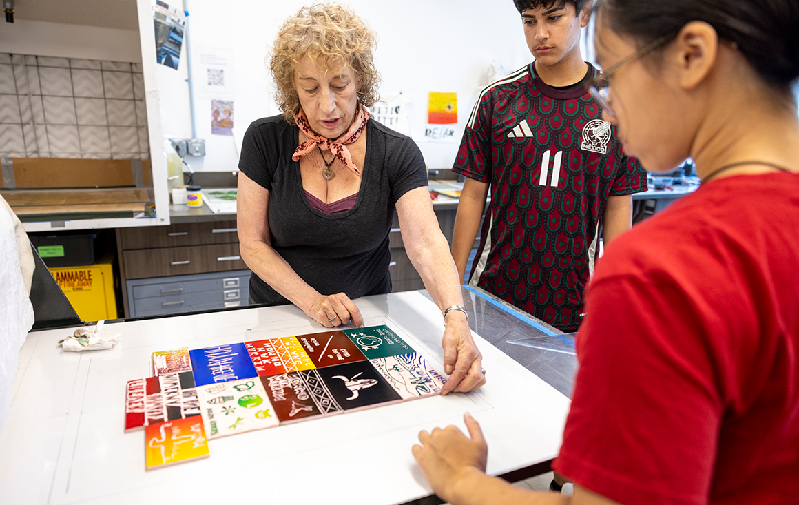 Woman in black shirt guides a linocut printing activity on a white table as two students stand by.