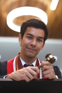 Young man with brown hair wears a suit with a red sash around his shoulder and gavel in his hand. On the ceiling behind him is a circular light. 