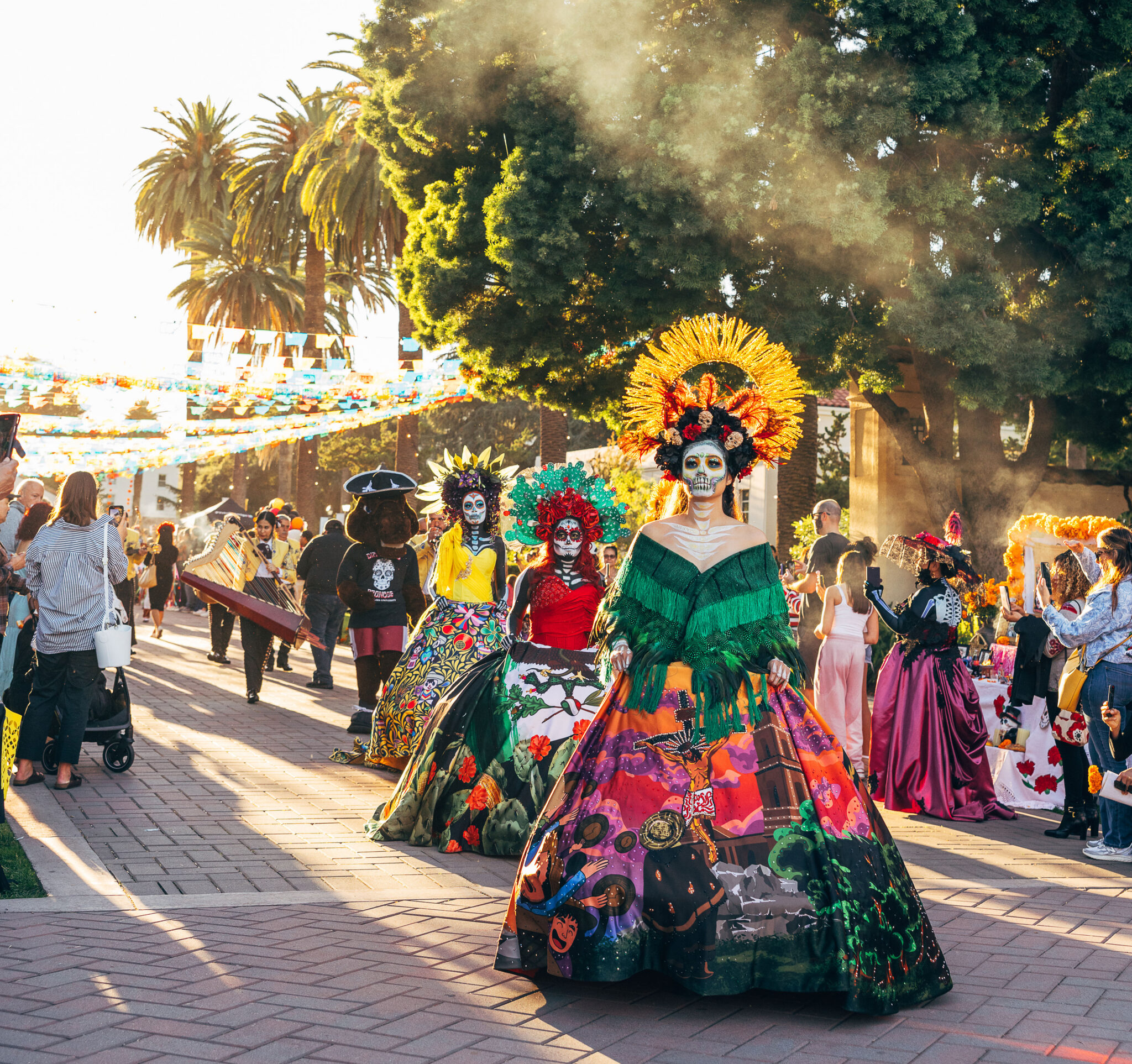women in colorful, elaborate dresses stand in a courtyard with festival-goers surrounding them