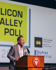 James Glaser stands at a podium in front of a projector screen with an event welcome.